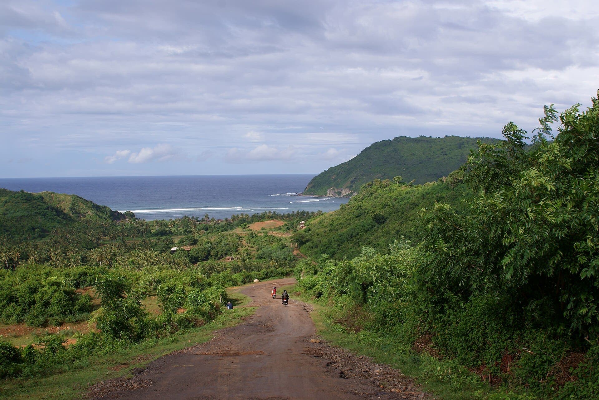 Two riders on the road down through green hills to the sea at Mawun, South Lombok
